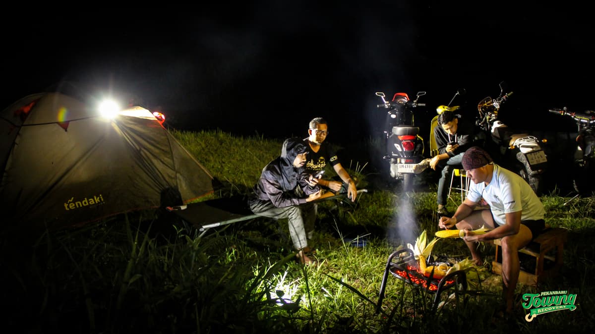Suasana camping santai di perbukitan bersama, dikelilingi tenda, motor, dan cahaya yang hangat.