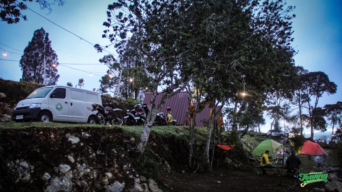 Suasana camping santai di perbukitan bersama, dikelilingi tenda, motor, dan cahaya yang hangat.